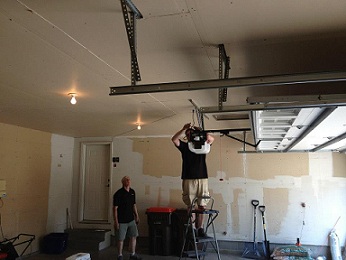Elite Garage Door technician with a black shirt and tan shorts standing on a ladder working on a garage door opener.