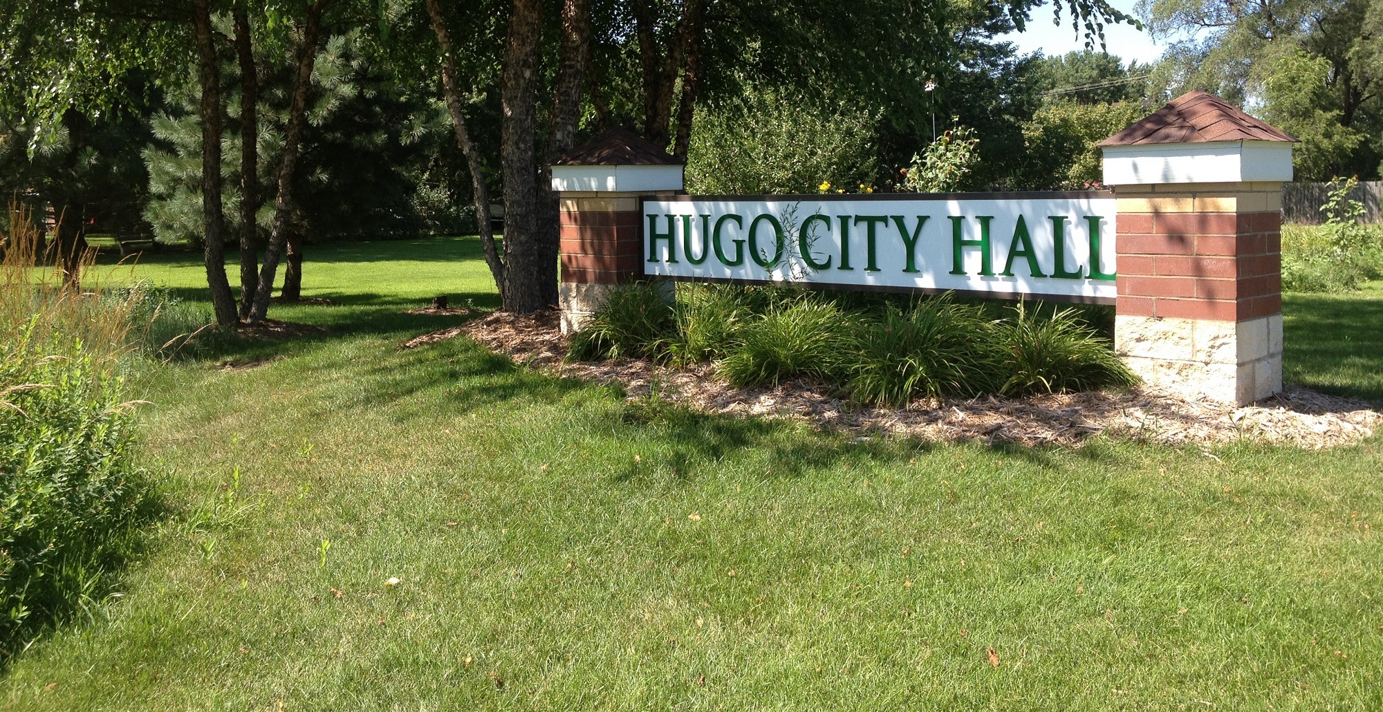 Hugo city hall sign with brick and signs and with bushes below the sign on a green lawn with trees in the background.