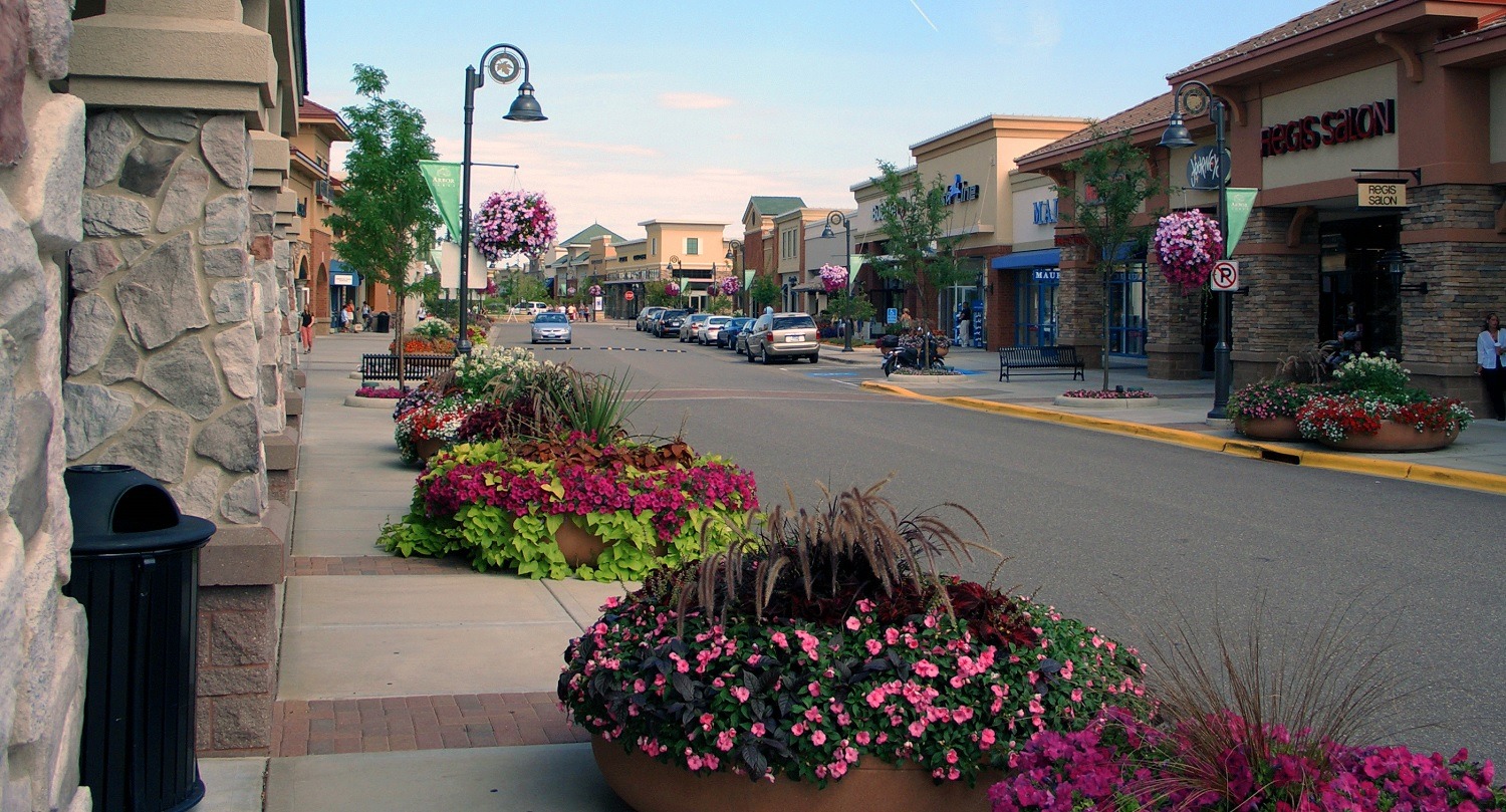 Street view of the arbor lakes outdoor mall with pink flowers and a red and green brushes with storefronts in the background.