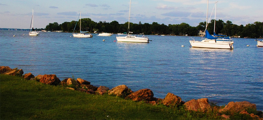 green grass and rocks in front of Lake Minnetonka with sail boats on a clear day with trees in the background.