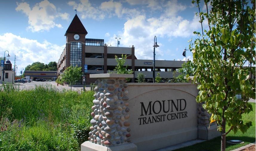 Rock and cement sign of Mound Transit Center with a parking ramp in the background with a cloudy sky behind the parking ramp.