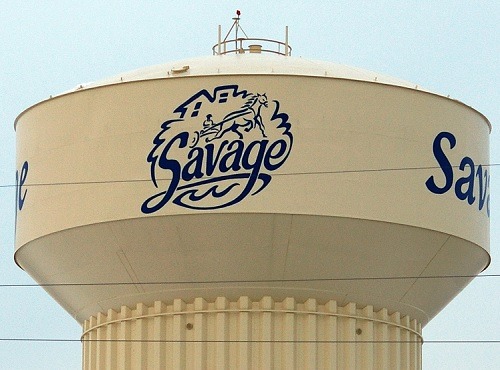 Off white water tower with blue savage lettering with a horse and carriage logo with a blue sky.
